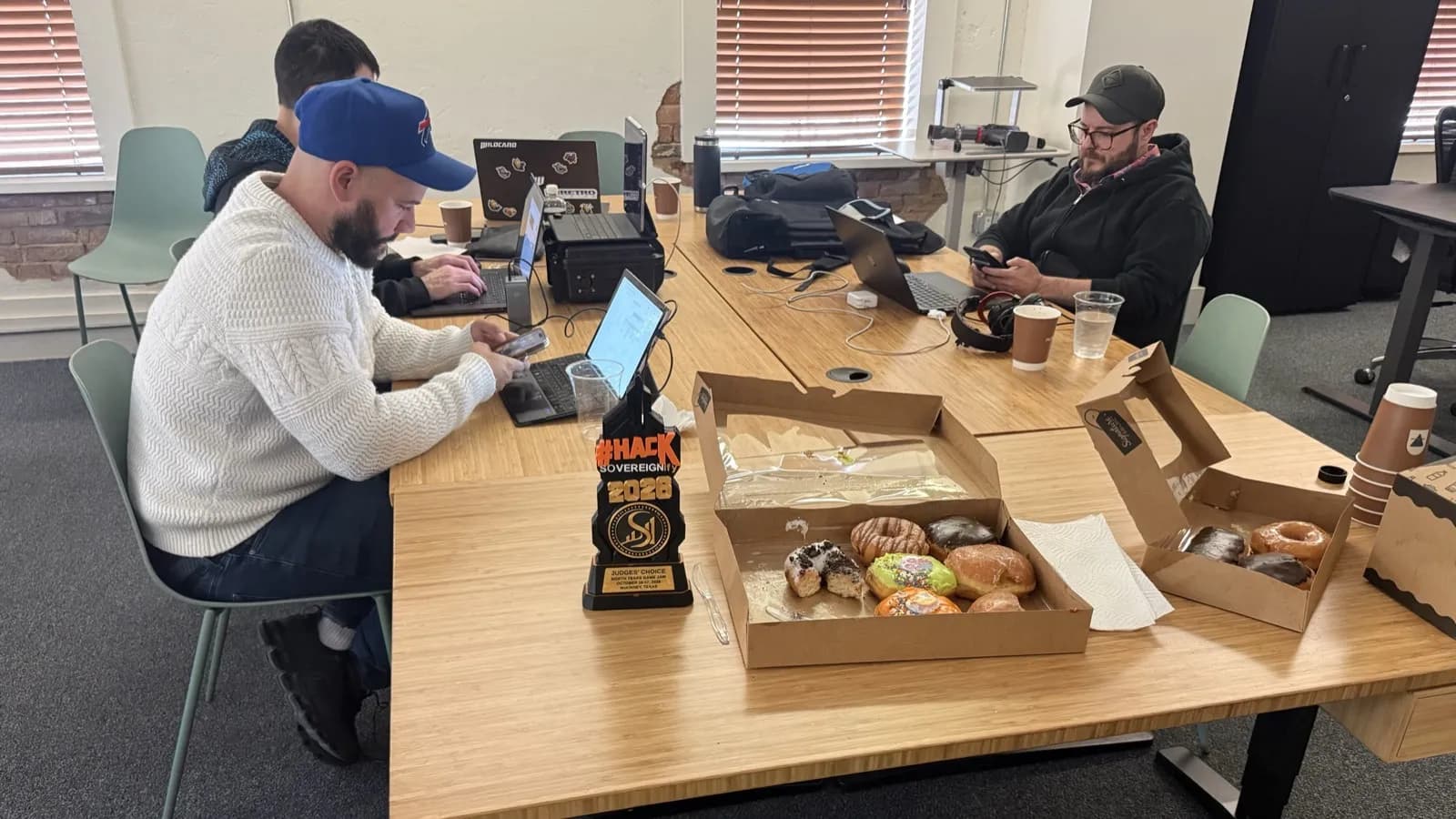 Three people at laptops around a long wooden table. In the foreground, the black #HACK × SOVEREIGNify 2026 Judges' Choice trophy stands beside open boxes of donuts.